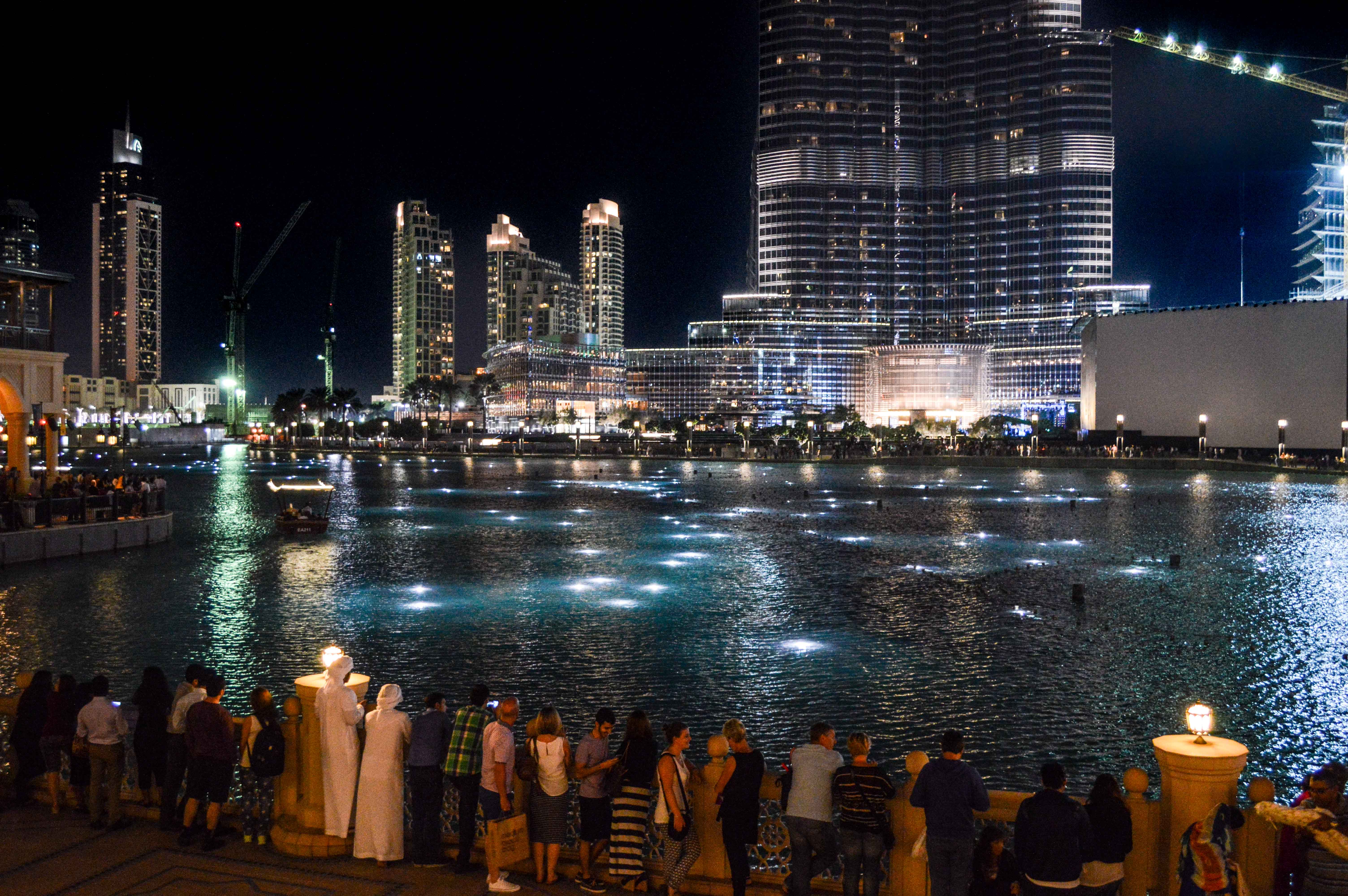 The image depicts a vibrant night scene in a modern city, likely Dubai, with tall skyscrapers illuminated against the night sky. People are gathered along the edge of a large water body, possibly a fountain, enjoying the view. The water is lit with numerous small lights, creating a reflective and serene atmosphere. The scene captures the essence of urban life and architectural grandeur.