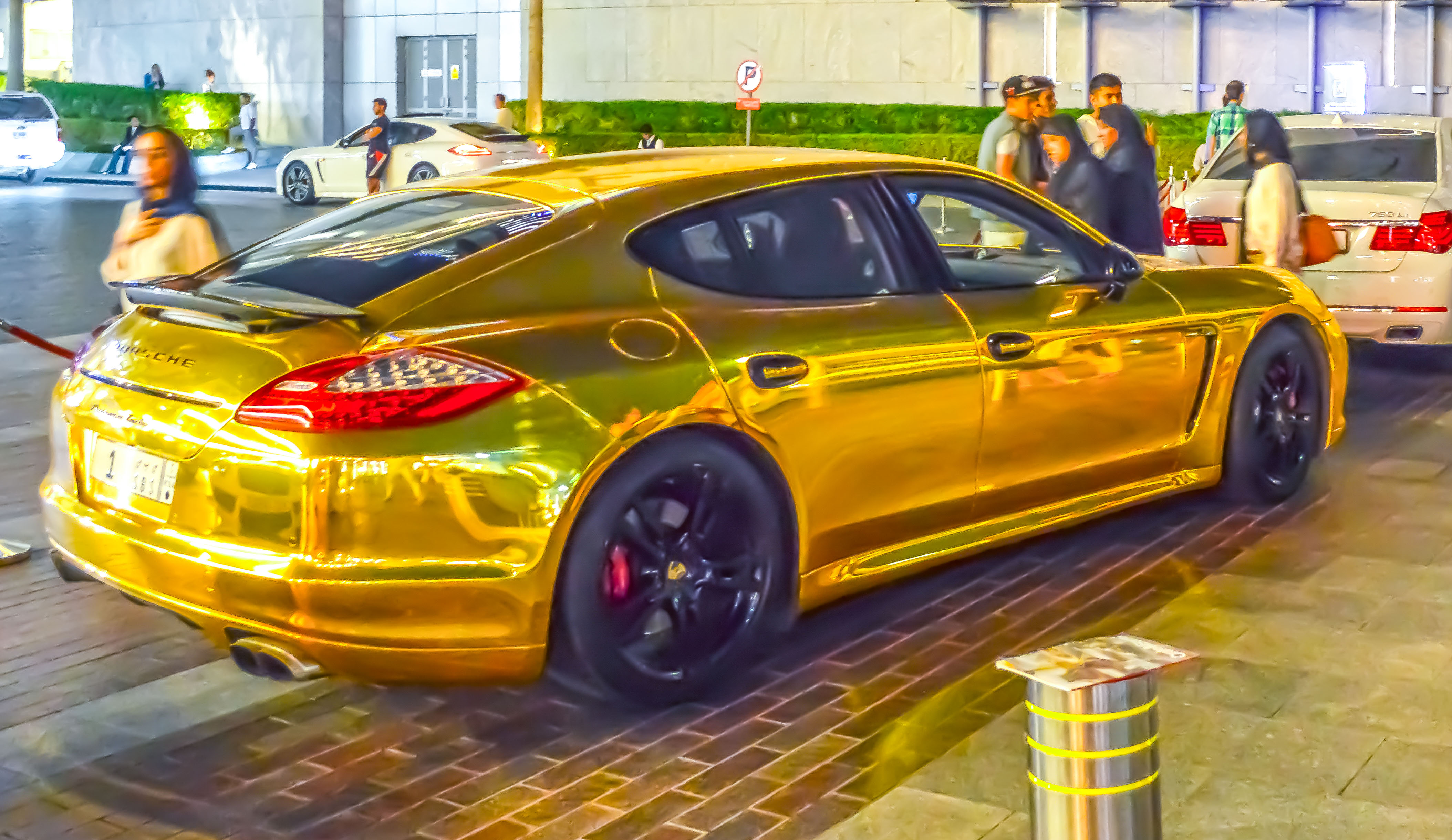 The image shows a shiny gold Porsche Panamera parked in a lot with people around it. The car has a distinctive and luxurious appearance, with a glossy finish and custom wheels. The setting appears to be an urban area, possibly near a commercial or residential building.