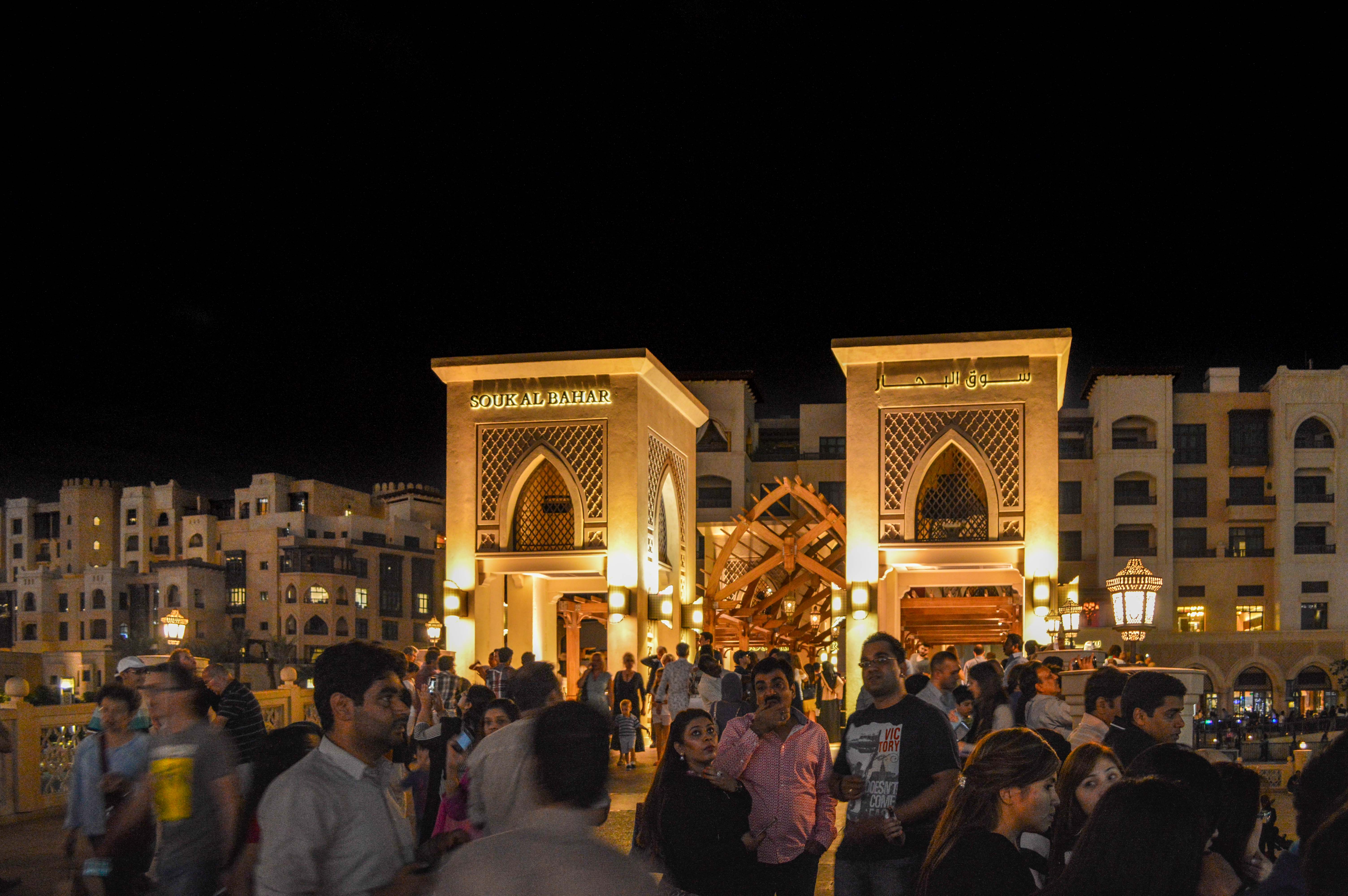 The image depicts a bustling night scene at the entrance of Souk Al Bahar, a market located in Dubai, UAE. The entrance is architecturally grand, illuminated with warm lights, and features traditional Arabic design elements. A large crowd of people is gathered in front of the entrance, suggesting a lively and popular destination. The surrounding buildings are also lit up, contributing to the vibrant atmosphere.
