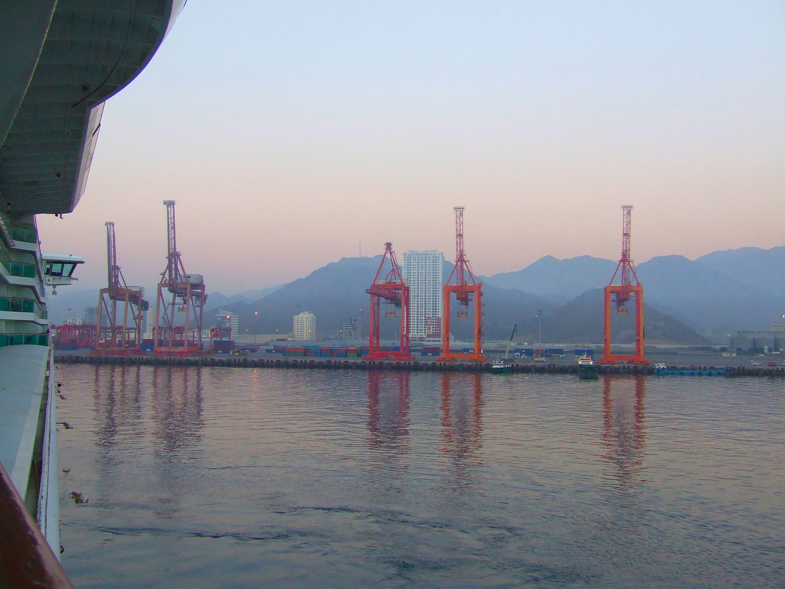 The image depicts a harbor scene at dusk or dawn, featuring several large red cranes used for loading and unloading shipping containers. The cranes are reflected in the calm water, with mountains and city buildings in the background. The scene is captured from a vantage point on a bridge or similar structure, providing a clear view of the harbor activities and the serene surroundings.