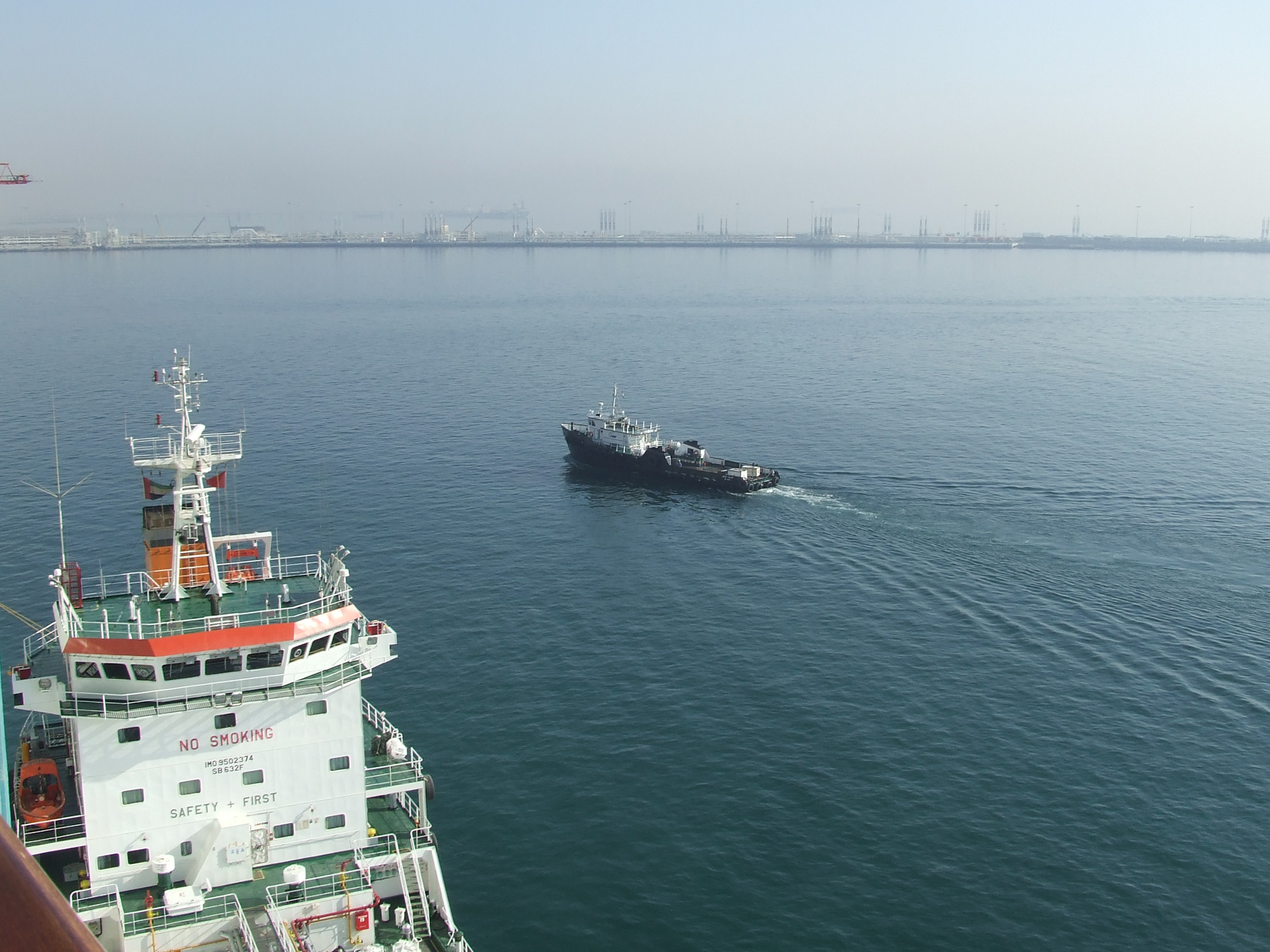 The image shows a maritime scene with two vessels on the water. The foreground features the upper deck of a large ship with visible safety markings such as 'NO SMOKING' and 'SAFETY FIRST'. In the background, a smaller vessel is seen navigating the water, heading towards a distant port with numerous cranes and industrial structures visible on the horizon.