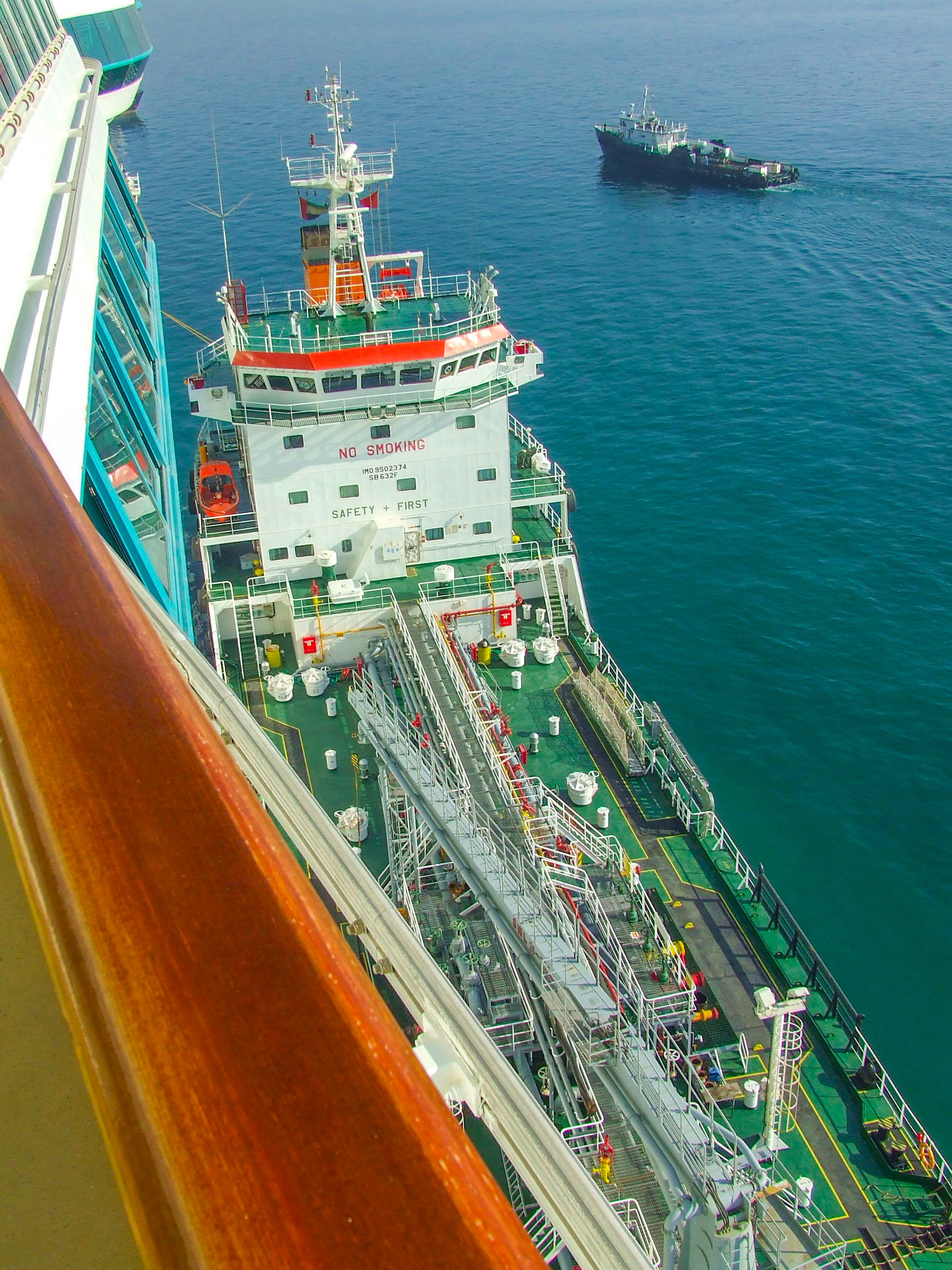The image depicts a harbor scene at dusk or dawn, featuring several large red cranes used for loading and unloading shipping containers. The cranes are reflected in the calm water, with mountains and city buildings in the background. The scene is captured from a vantage point on a bridge or similar structure, providing a clear view of the harbor activities and the serene surroundings.