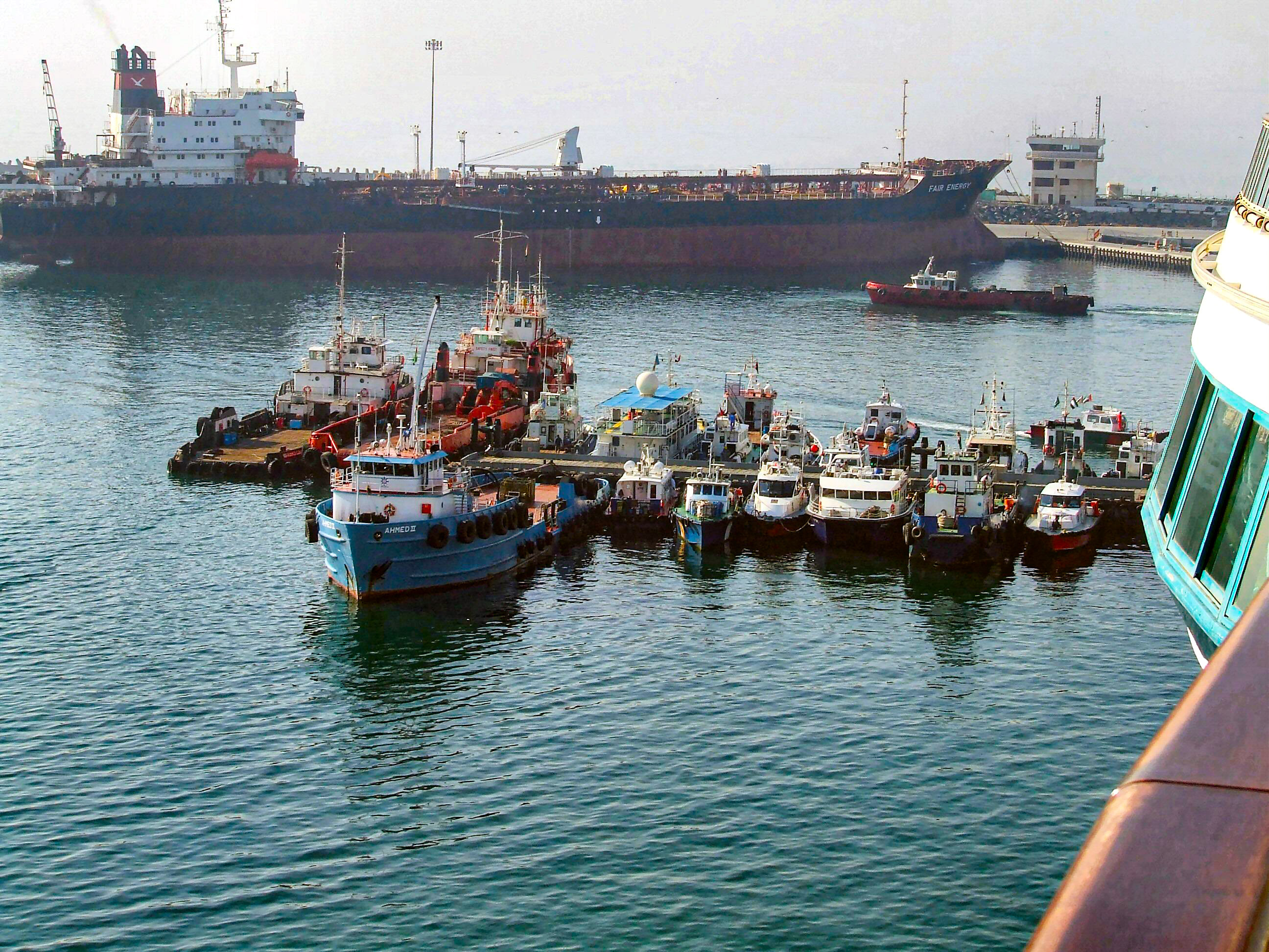The image shows a busy harbor scene with numerous small boats and tugboats docked in the foreground. In the background, there are larger vessels, including a significant cargo ship named 'FAIR ENERGY.' The harbor appears to be a bustling port with various maritime activities.