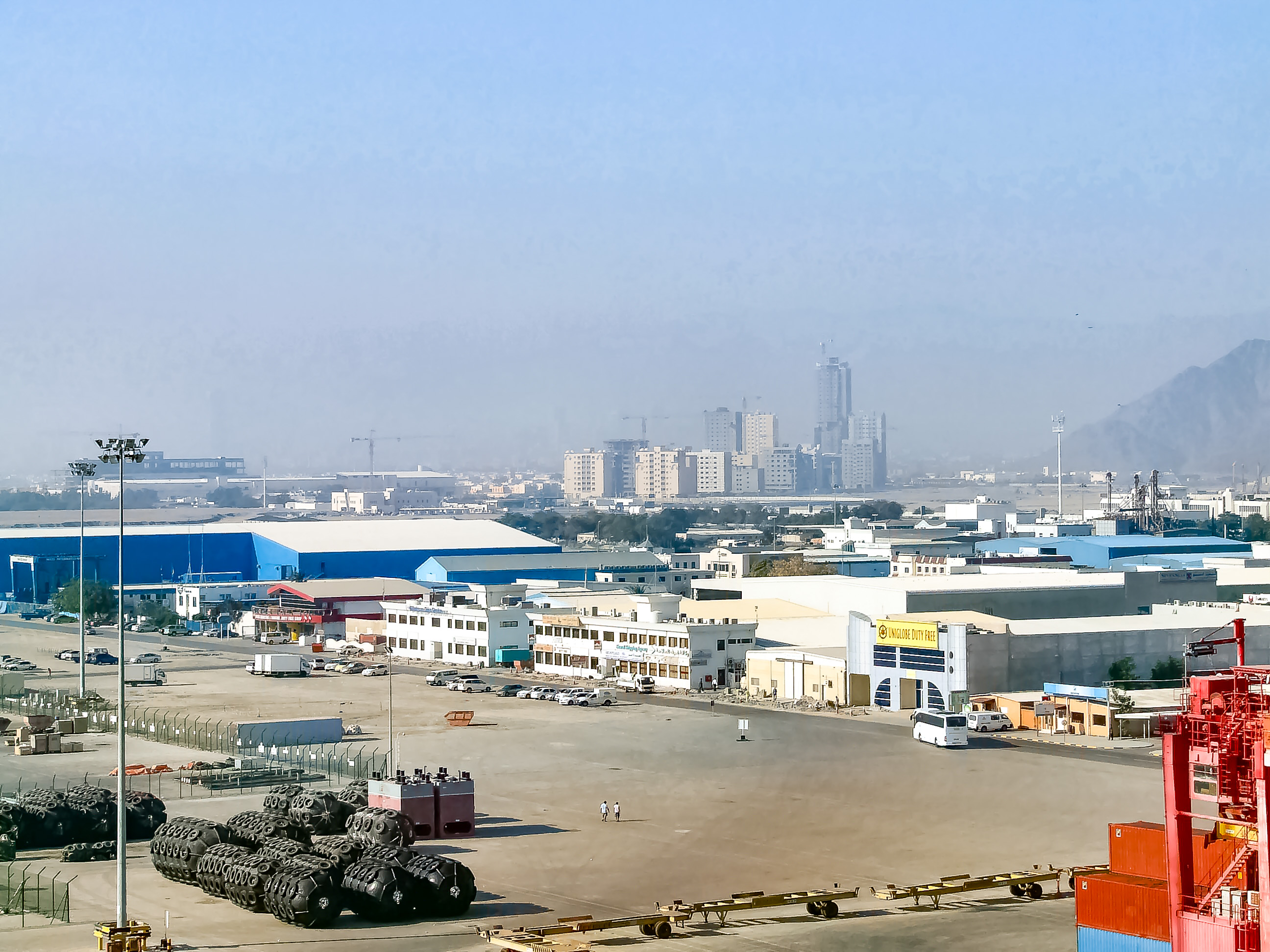 The image shows an industrial area with warehouses, buildings, and a port facility. In the foreground, there are stacks of tires and various vehicles, including cars and buses. The background features a city skyline with several high-rise buildings and construction cranes, indicating ongoing development. The sky is hazy, suggesting the presence of dust or smog.