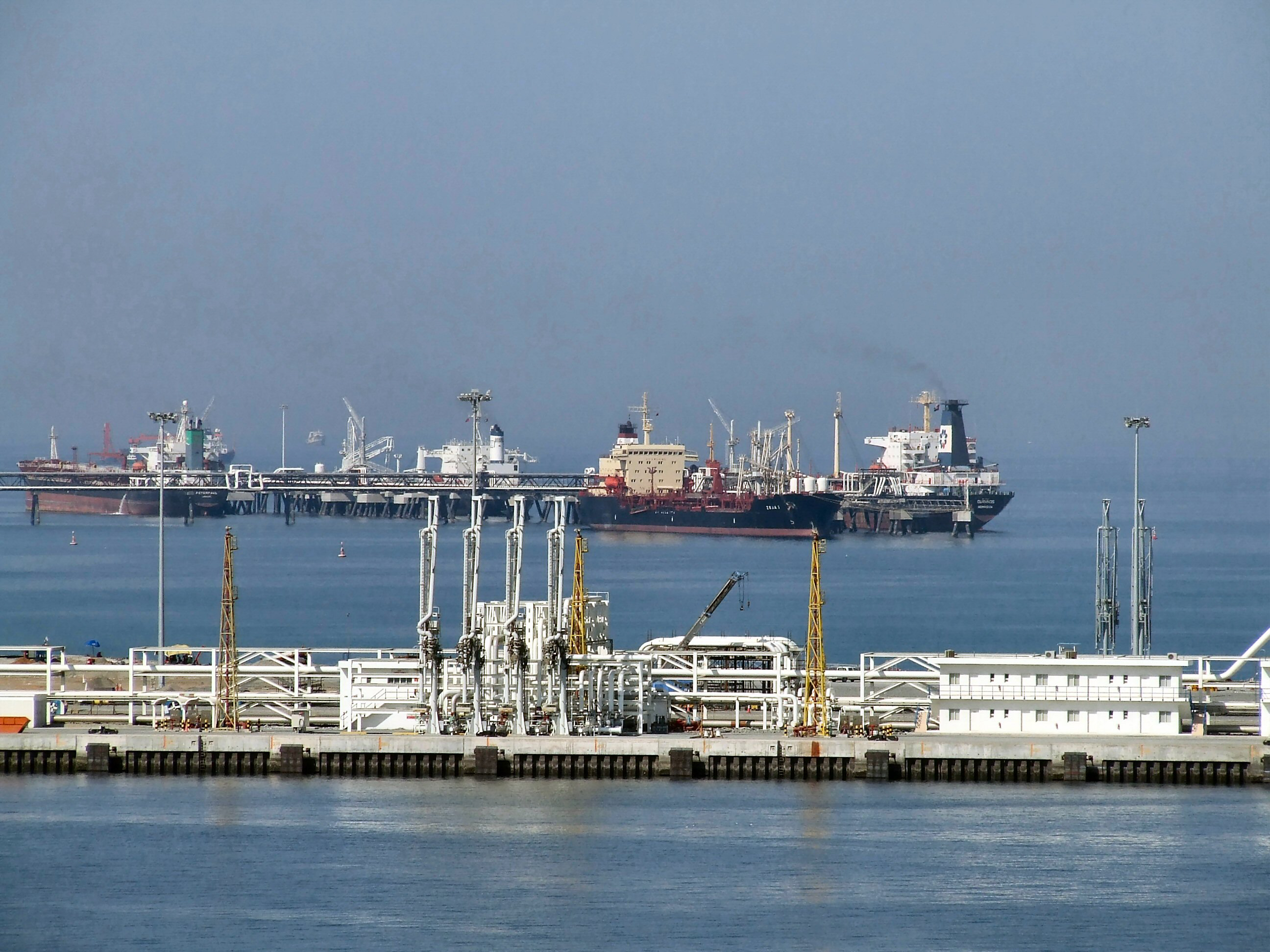 The image depicts a bustling port area with multiple large ships docked at a pier. The foreground shows industrial infrastructure, including pipelines, cranes, and various buildings. The background features more ships and port facilities, with a hazy sky overhead. The water in the foreground is calm, reflecting the industrial structures along the shore.