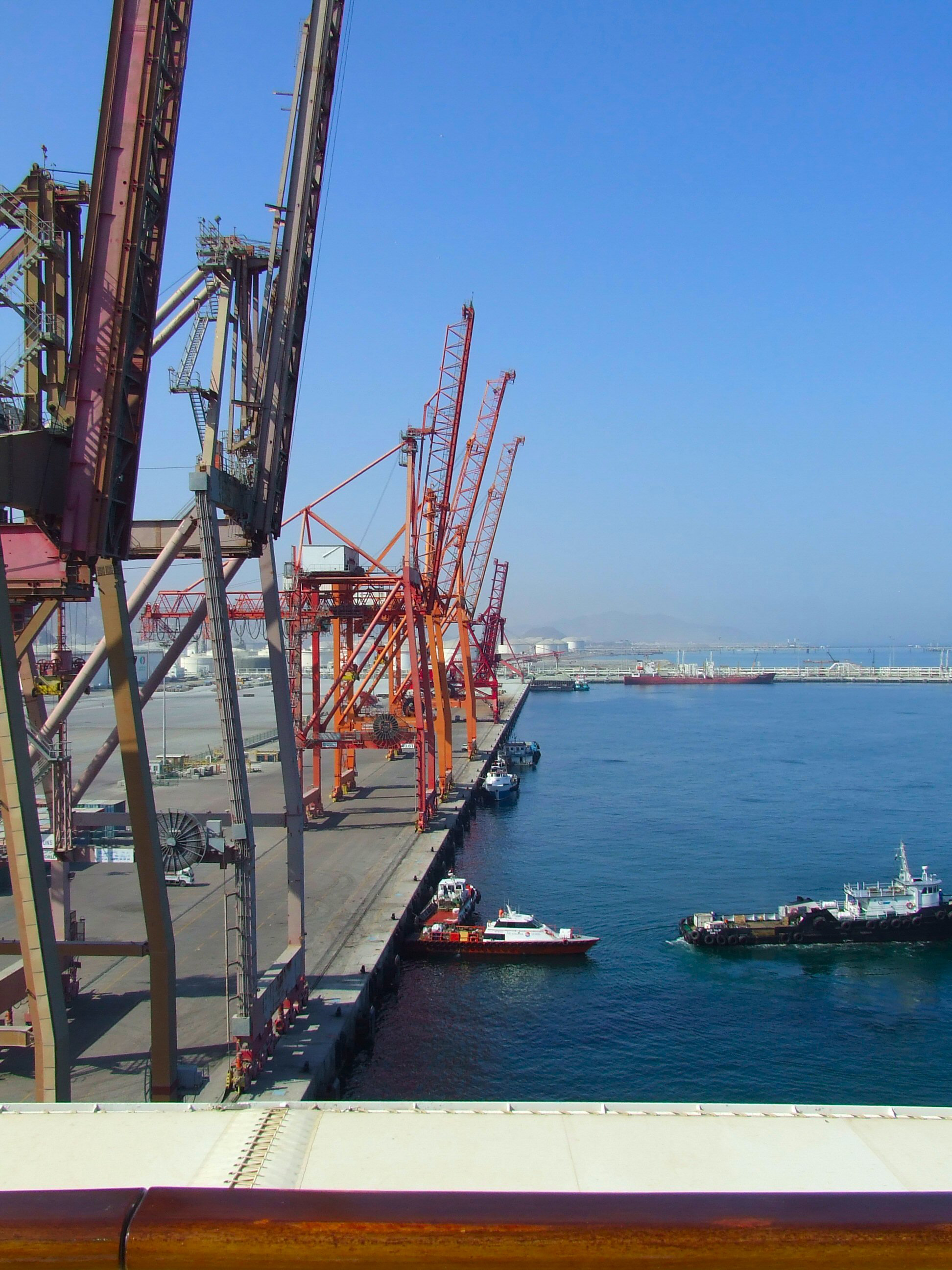 The image depicts a bustling port area with numerous cranes and docks. Several boats and ships are visible in the water, and the scene is set against a clear blue sky. The cranes, painted in red and yellow, are positioned along the docks, ready for loading and unloading activities. The water is calm, and the overall atmosphere suggests a busy, yet organized, maritime environment.