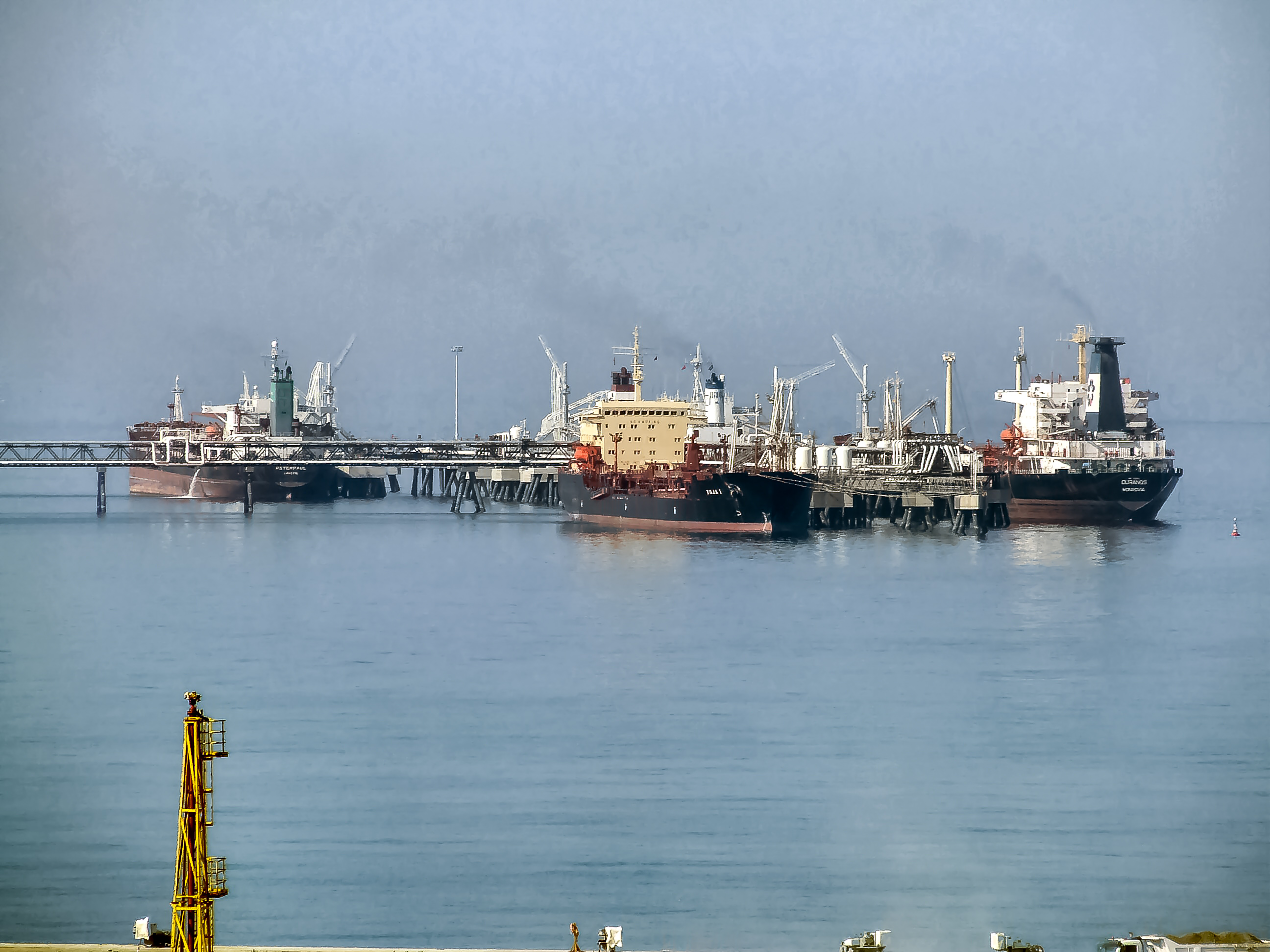 The image shows a port with several large ships docked. The ships appear to be involved in industrial activities, possibly related to oil or chemical transport. The weather seems hazy, and the water is calm. There are structures and equipment on the ships, indicating they are equipped for specific industrial purposes. The port infrastructure includes a pier and various support facilities.