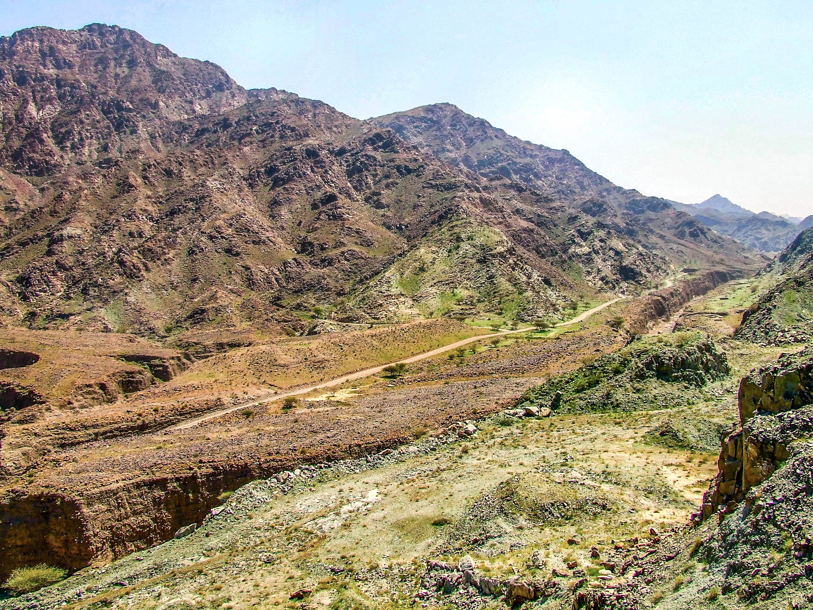 The image depicts a rugged, mountainous landscape with steep, rocky slopes and sparse vegetation. A narrow, winding dirt path can be seen traversing the terrain, leading up towards the higher elevations. The overall scene is arid, with patches of green shrubs and grass interspersed among the rocky outcrops. The sky above is clear, suggesting a sunny day. The terrain appears challenging and remote, suitable for hiking or exploration in a natural, untouched environment.
