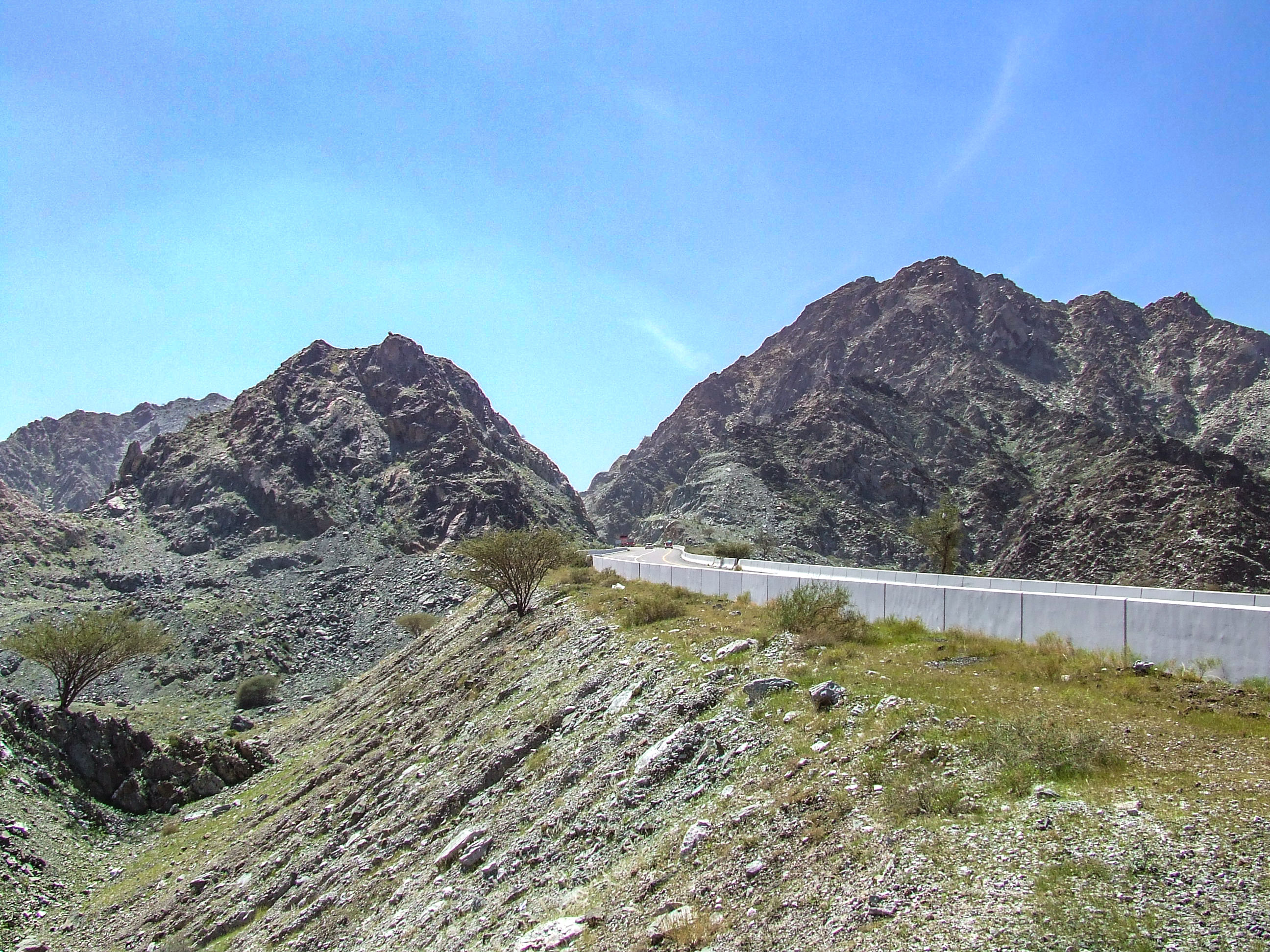 The image depicts a rugged mountainous landscape with two prominent peaks under a clear blue sky. In the foreground, there is a sloping terrain with sparse vegetation and a few small trees. A white wall or barrier runs horizontally across the middle of the image, possibly indicating a road or a boundary.