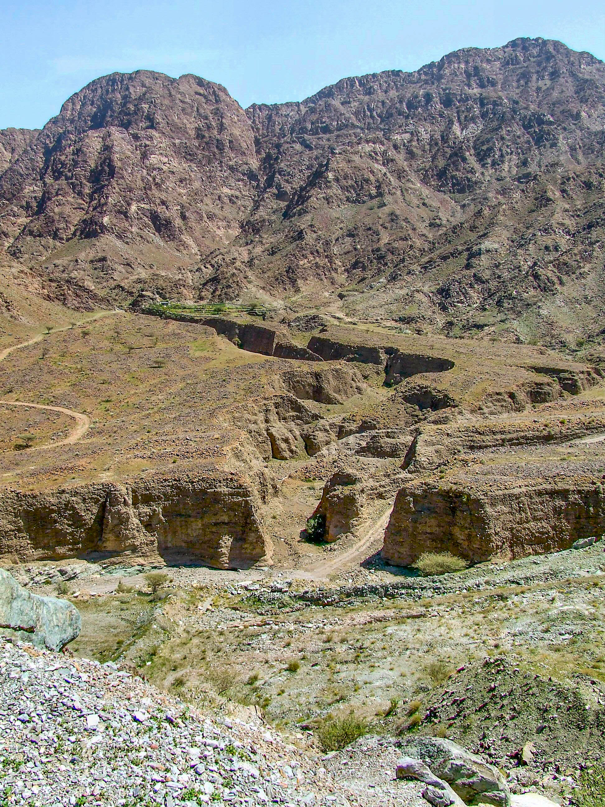 The image depicts a rugged, mountainous landscape with steep, rocky terrain and sparse vegetation. The foreground features eroded soil and small plants, while the background showcases towering, barren mountains under a clear sky.