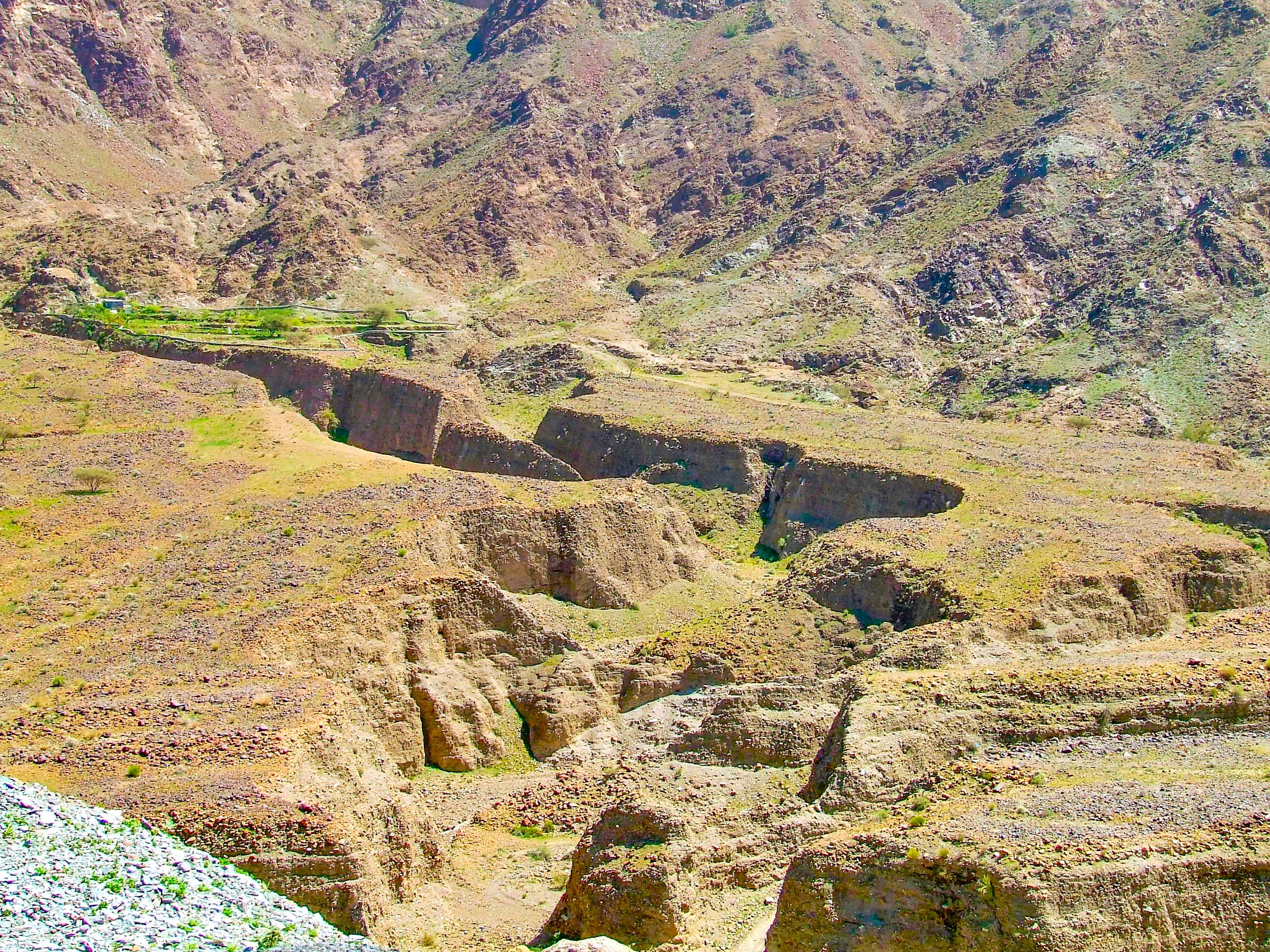 The image depicts an ancient archaeological site with visible ruins and remnants of structures built into a hillside. The terrain is rugged and uneven, with patches of grass and vegetation scattered across the rocky surface. The ruins appear to be made of stone and are partially eroded, indicating significant age. The site is likely of historical or cultural importance, possibly an ancient settlement or ceremonial site.