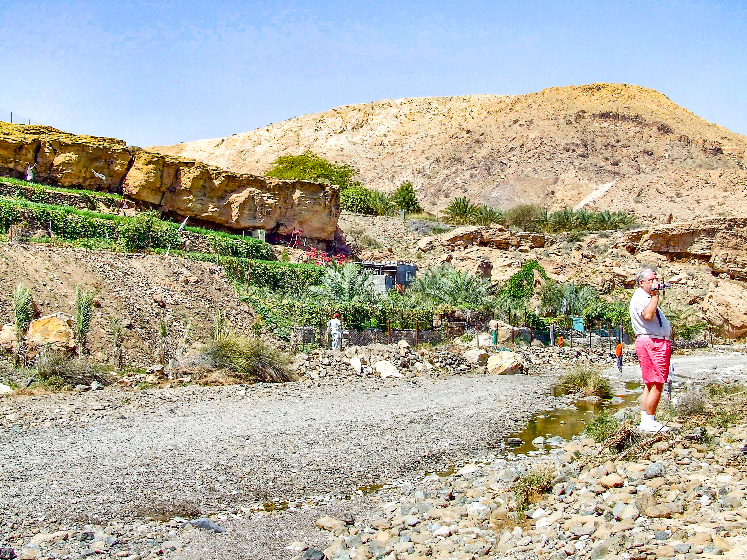 The image depicts a man standing in a rocky, arid landscape with a small stream of water in the foreground. He is dressed in casual clothing, including a white shirt, red shorts, and white shoes, and is holding a camera or binoculars. The background features a rocky hillside with sparse vegetation, including small trees and bushes. There are also some structures, possibly small buildings or shelters, partially obscured by the vegetation. The sky is clear and blue, indicating a sunny day.