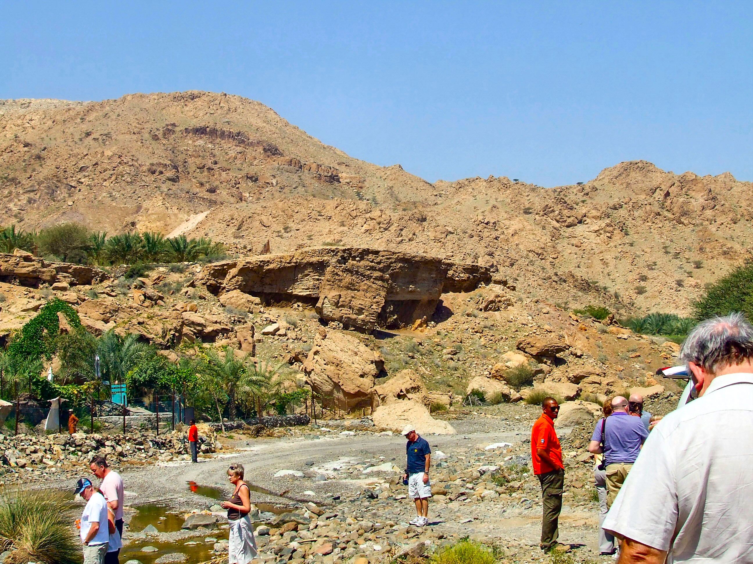 The image depicts a group of people exploring a rocky, arid landscape with a prominent rocky hill in the background. The area appears to be a historical or archaeological site, with some greenery and small structures visible. The people seem to be on a guided tour, with one person in a red shirt likely serving as the guide.