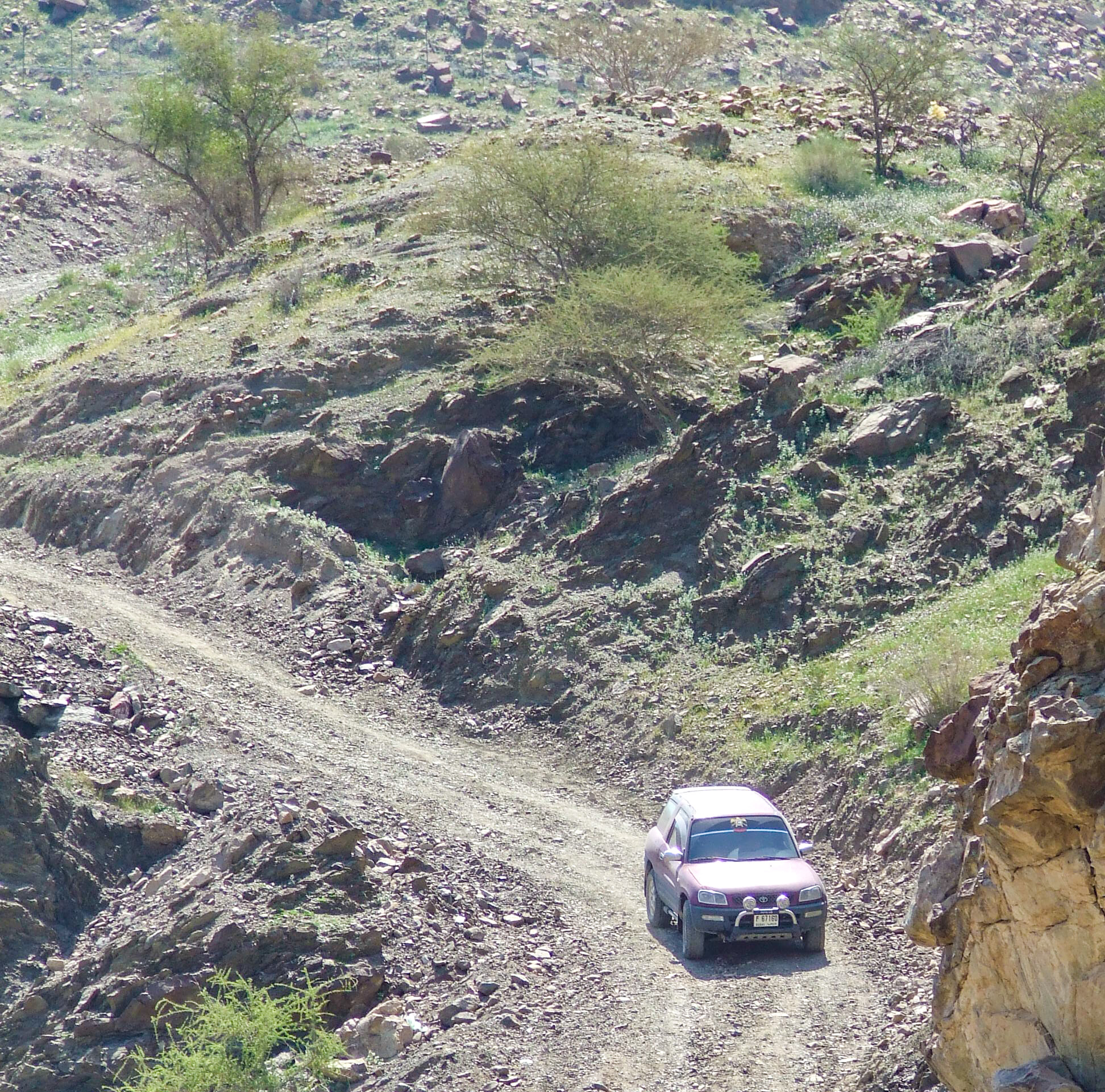 The image depicts a red SUV driving on a rugged, rocky trail in a mountainous area. The terrain is uneven with loose rocks and sparse vegetation. The vehicle appears to be navigating the challenging path, suggesting an off-road adventure.